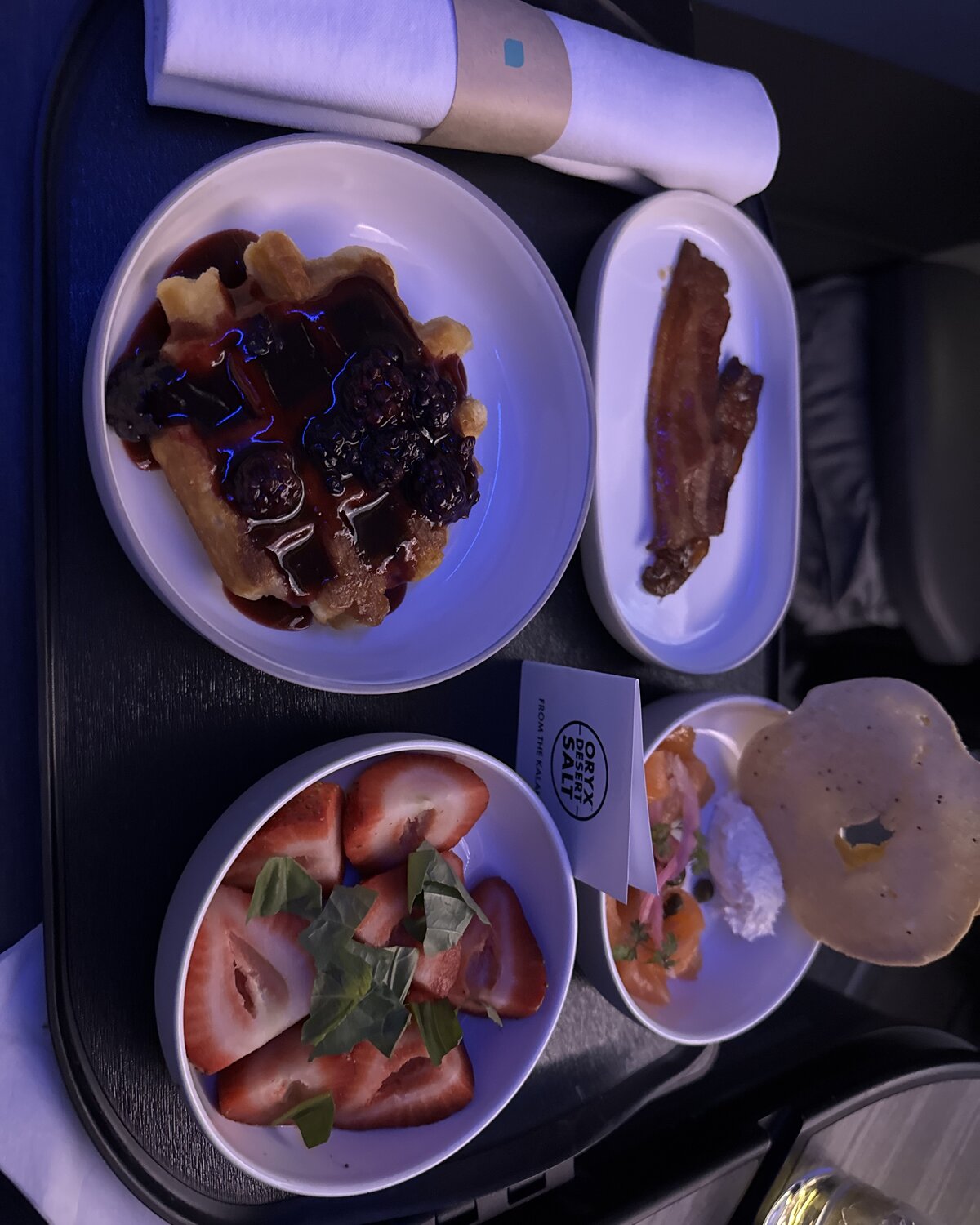 JetBlue Mint breakfast tray — waffle with blackberry preserves and berries, fresh strawberries with basil, smoked salmon with bagel crostini, and a side of maple bacon